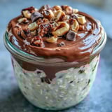 Spoon dipping into jars of chilled Peanut Butter Cup Overnight Oats, showing a creamy texture and a topping of mini chocolate chips.