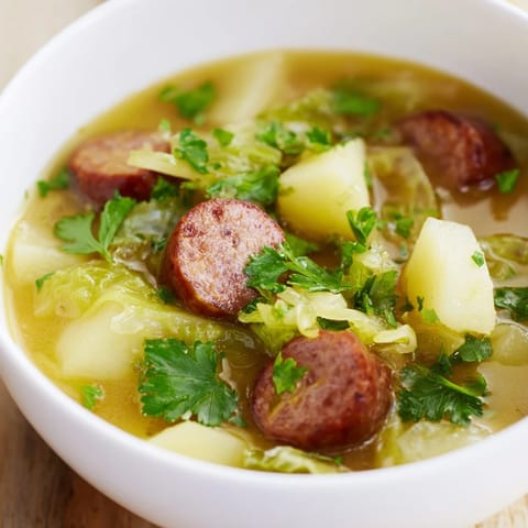 Hearty Sausage, Potato and Cabbage Soup served hot in a mug with crusty bread on a wooden table.
