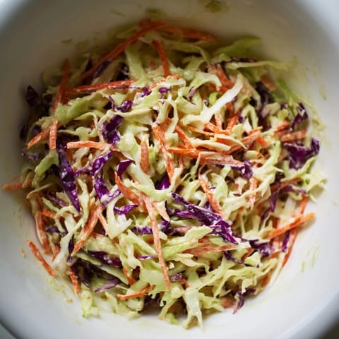 Freshly tossed Coleslaw With Creamy Dressing piled high in a white bowl, garnished with a sprinkle of celery seed beside grilled chicken.